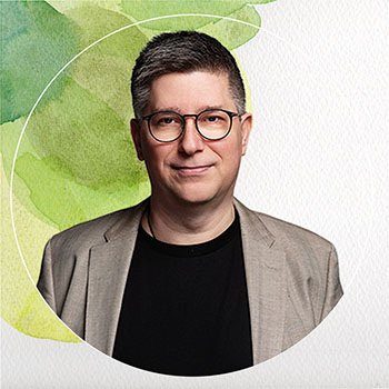 A middle aged white man with short brown hair. He wears glasses with a thing oval black rim. He is wearing a black t-shirt with a tan sports coat.